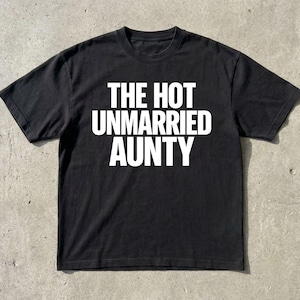 May include: Black t-shirt with the white text "THE HOT UNMARRIED AUNTY". The shirt has short sleeves and a crew neck. The shirt is displayed on a gray surface.