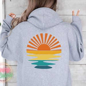 May include: A gray hooded sweatshirt with a retro-style sunset graphic on the back. The graphic features a yellow, orange, and red sun with rays, reflected in a blue and green body of water.