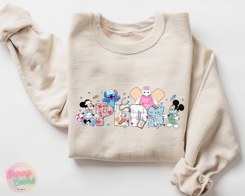 May include: A beige sweatshirt with a graphic print of the word "FRIENDS" spelled out with cartoon characters. The characters are Mickey Mouse, Stitch, and Baymax, all dressed as nurses. The letters are decorated with medical symbols and designs.