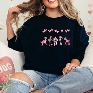 May include: A black sweatshirt with a pink and white design featuring characters from the Toy Story movie. The characters are lined up in a row and include Woody, Buzz Lightyear, Jessie, Rex, and Bullseye. There are pink Mickey Mouse ears above the characters.