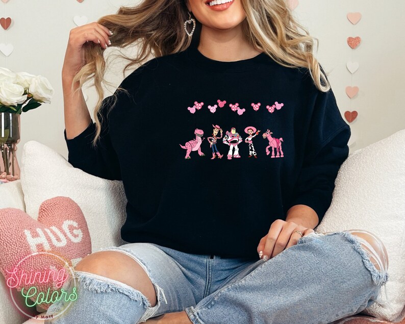May include: A black sweatshirt with a pink and white design featuring characters from the Toy Story movie. The characters are lined up in a row and include Woody, Buzz Lightyear, Jessie, Rex, and Bullseye. There are pink Mickey Mouse ears above the characters.