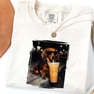 May include: Cream-colored t-shirt featuring a graphic of a dachshund wearing sunglasses and drinking iced coffee. The dog is in a black and white framed image. A gold necklace and a brown hat are also visible.