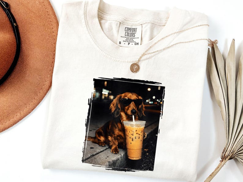 May include: Cream-colored t-shirt featuring a graphic of a dachshund wearing sunglasses and drinking iced coffee. The dog is in a black and white framed image. A gold necklace and a brown hat are also visible.