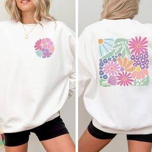May include: White sweatshirt with a floral design on the front and back. The flowers are in shades of pink, blue, yellow, and green.