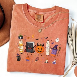 May include: A terracotta-colored t-shirt featuring a graphic print of various Halloween-themed characters including a mummy pencil, a vampire book, a pumpkin with horns, a mummy crayon, and a purple eraser with a witch hat and broomstick.