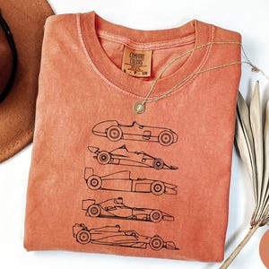 K&ouml;nnte beinhalten: Korallenfarbenes Comfort Colors T-Shirt mit einer schwarzen Strichzeichnung von f&uuml;nf Oldtimer-Rennwagen. Das Shirt hat einen Rundhalsausschnitt und ein kleines Etikett. Eine goldene Halskette und ein brauner Hut liegen neben dem Shirt.