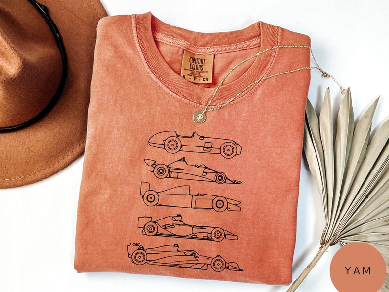 K&ouml;nnte beinhalten: Korallenfarbenes Comfort Colors T-Shirt mit einer schwarzen Strichzeichnung von f&uuml;nf Oldtimer-Rennwagen. Das Shirt hat einen Rundhalsausschnitt und ein kleines Etikett. Eine goldene Halskette und ein brauner Hut liegen neben dem Shirt.
