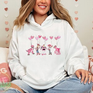 May include: White hoodie with a pink and white graphic of the Toy Story characters holding pink balloons with white polka dots. The characters include Woody, Buzz Lightyear, Jessie, Rex, and Bullseye.