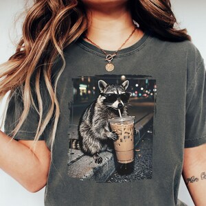 May include: Dark grey t-shirt featuring a raccoon wearing sunglasses and drinking iced coffee. The raccoon is depicted in a nighttime urban setting. The shirt is a comfortable, casual style.