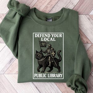 May include: Olive green sweatshirt with the text "DEFEND YOUR LOCAL PUBLIC LIBRARY." The graphic depicts a knight on a black cat holding a flag. The sweatshirt has long sleeves and a crew neck.