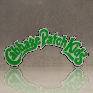 Può includere: Un logo verde e bianco dei Cabbage Patch Kids. Le parole "Cabbage Patch Kids" sono in un font stilizzato con un aspetto strutturato e puntinato. Il logo si riflette su una superficie lucida.