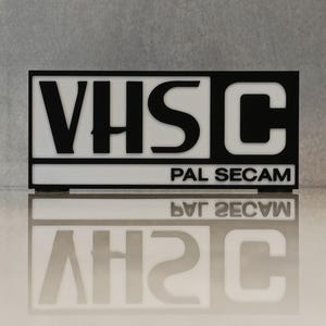 Peut inclure: Un panneau rectangulaire noir et blanc avec le texte "VHS C" en lettres majuscules grasses. En dessous, on peut lire "PAL SECAM". Le panneau se reflète sur une surface brillante.