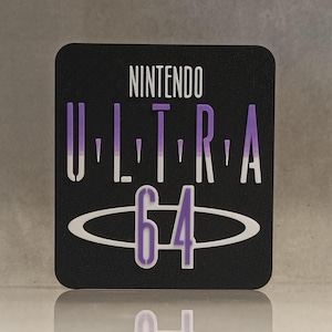 May include: A black square with the words "NINTENDO ULTRA 64" in white and purple. The text is arranged vertically, with "ULTRA" in a stylized font and "64" inside an oval. The image is reflected on a shiny surface.