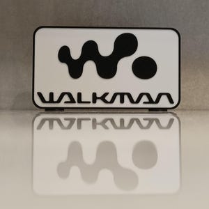 May include: A white rectangular object featuring a black logo and the word "WALKMAN" in black. The logo is a stylized "W" shape and a circle. The object is reflected on a glossy surface.