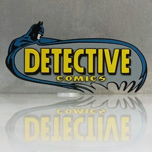 Peut inclure: Un panneau sur le thème de Batman avec les mots "DETECTIVE COMICS" en jaune, bordés de noir, sur un fond gris. Le panneau a une bordure bleue et une silhouette de Batman. Le reflet du panneau est visible en dessous.