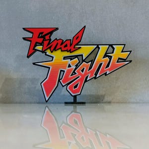 May include: A colorful "Final Fight" logo, featuring red, yellow, and orange gradients with a black outline. The logo is mounted on a small black stand, casting a reflection on a glossy surface. The design is reminiscent of a classic video game.