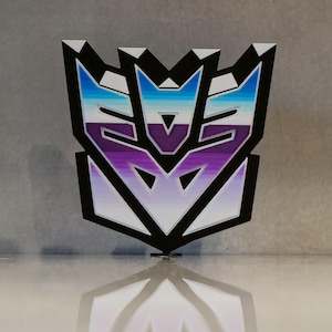 Könnte beinhalten: Ein 3D-gedrucktes Emblem mit schwarzem Umriss und einem Farbverlauf aus Blau, Lila und Weiß. Das Emblem ist ein bekanntes Symbol aus dem Transformers-Franchise, mit einer Spiegelung darunter.