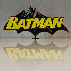 May include: A Batman logo sign with the word "BATMAN" in yellow letters with a red outline, set against a black bat-shaped background. The image includes a teal-coloured Batman silhouette. The sign is reflected below.