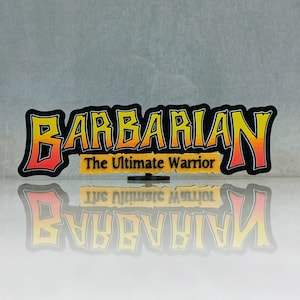 May include: A graphic with the word "BARBARIAN" in yellow and red, outlined in black, with the text "The Ultimate Warrior" below. The graphic is reflected on a shiny surface.