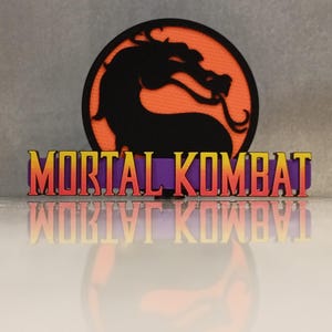 May include: A black dragon silhouette on an orange circle, with the words "MORTAL KOMBAT" in yellow, orange, and purple. The logo is reflected below. The image is a close-up.