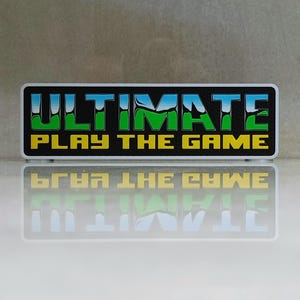 Op de afbeelding: Rechthoekige sticker met de tekst "ULTIMATE PLAY THE GAME" in een gedurfd, kleurrijk ontwerp. De tekst is in groen, blauw en geel, met een zwarte rand. De sticker wordt weerspiegeld op een wit oppervlak.