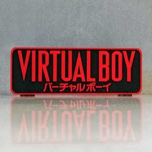 May include: A rectangular black and red sign with the words "VIRTUAL BOY" in bold red letters. Below the English text is Japanese text. The sign is reflected in the surface below.