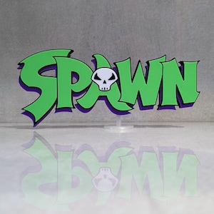 Op de afbeelding: Een driedimensionaal bord met het woord SPAWN in een gestileerd groen lettertype met een paarse omlijning. De letter A is vervangen door een wit schedelpictogram met zwarte oogkassen en een kleine zwarte neus. Het bord staat op een standaard.