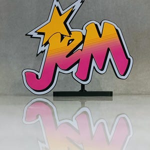 May include: A colorful JM logo in a retro style. The design features a yellow star and gradient lettering in yellow and pink, outlined in black. The logo is mounted on a black stand.