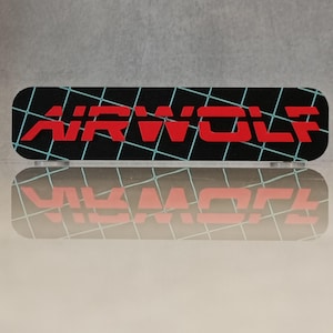 May include: A black, rectangular object featuring the word "AIRWOLF" in red, with a teal grid pattern. The object's reflection is visible on a clear surface below. The design is reminiscent of retro graphics.
