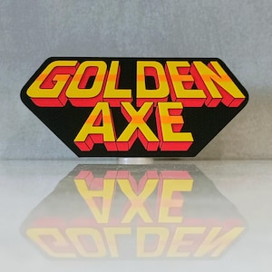 May include: A black, retro-style patch with the words "GOLDEN AXE" in yellow, orange, and red 3D lettering. The patch has a unique hexagonal shape and is reflected on a glossy surface.