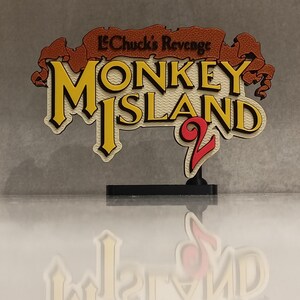 May include: A 3D-printed sign for "Monkey Island 2: LeChuck's Revenge" in yellow, cream, red, and brown. The sign is mounted on a black base, with a reflection visible below. The text is in a stylized font.