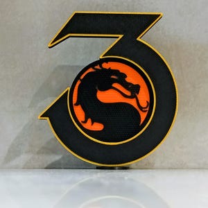 Könnte beinhalten: Ein dreidimensionales Schild in Form der Zahl Drei mit schwarzer Oberfläche und goldenen Kanten. In der Mitte der Zahl befindet sich ein kreisförmiges Emblem, das die Silhouette eines schwarzen Drachen auf leuchtend orangefarbenem Grund zeigt.