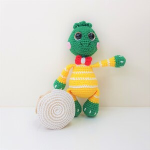 May include: A green and yellow crocheted dinosaur toy with a red bow tie. The dinosaur has black eyes and pink cheeks. The toy is standing next to a white crocheted object.