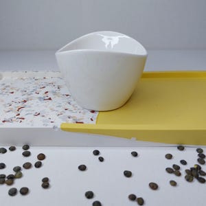 Könnte beinhalten: Eine weiße Keramikschale steht auf einem weiß-gelben Tablett. Das weiße Tablett hat ein gesprenkeltes Design mit roten, blauen und braunen Akzenten. Kleine, dunkle Samen sind um das Tablett verstreut.