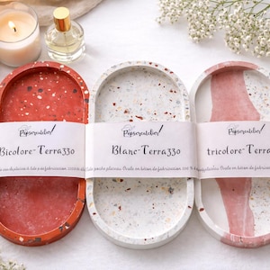 Könnte beinhalten: Drei ovale Terrazzo-Tabletts in Rot, Weiß und Rosa, jedes mit einem einzigartigen gesprenkelten Muster. Die Tabletts sind mit "Bicolore-Terra330", "Blanc-Terra330" und "tricolore-Terra330" beschriftet. Eine Kerze, eine Parfümflasche und Blumen sind im Hintergrund.