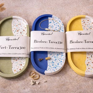 Può includere: Tre vassoi ovali in verde, blu e giallo, ciascuno con un design a terrazzo su una metà. I vassoi hanno il testo "Vert-Terra300" e "Bicolore-Terra330". Gioielli sono posti sul vassoio blu.