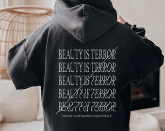 Puede incluir: Sudadera con capucha negra con el texto "BEAUTY IS TERROR" apilado, con una línea que atraviesa cada palabra. La cita "whatever we call beautiful, we quiver before it" está impresa debajo.