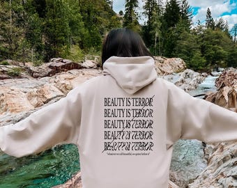 Puede incluir: Una sudadera con capucha beige con la frase "BEAUTY IS TERROR" repetida varias veces en la espalda. El texto es negro y la sudadera la lleva puesta una persona de pie en un entorno rocoso y natural.