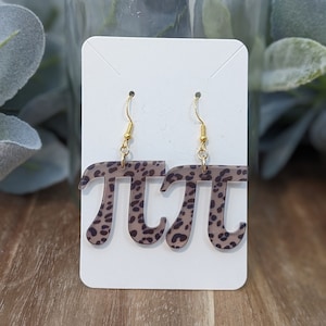 May include: A pair of dangle earrings featuring the mathematical symbol pi. The earrings have a tan and black leopard print pattern. They are suspended from gold-colored fish hook ear wires and displayed on a white card.