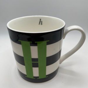 Puede incluir: Una taza de cerámica blanca con un patrón de rayas blancas y negras y una letra "H" verde en la parte delantera. La taza tiene un asa negra y la letra "h" está escrita en el interior de la taza.