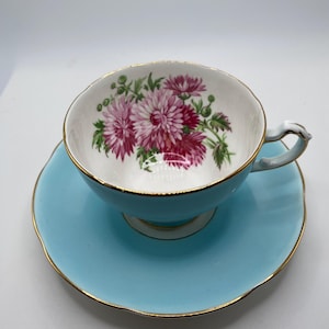Może przedstawiać: Zestaw filiżanki i spodka do herbaty w stylu vintage. Filiżanka i spodek są w kolorze jasnoniebieskim ze złotymi obramowaniami. Wnętrze filiżanki zdobi kwiatowy wzór z różowymi i fioletowymi kwiatami.