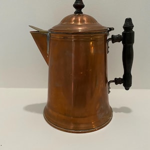 May include: A copper teapot with a wooden handle and a lid. The teapot has a shiny finish and a classic design.