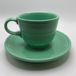 Puede incluir: Un juego de taza y platillo de té de cerámica verde. La taza tiene una forma redondeada y un asa. El platillo tiene un borde festoneado.