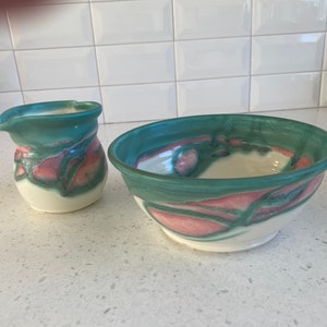 May include: A ceramic creamer and bowl set. The creamer and bowl are white with teal, pink, and green swirled designs. The bowl has a wide rim and a rounded shape. The creamer has a spout and a handle. The set is handmade.