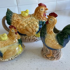 May include: A ceramic teapot, creamer and sugar bowl set featuring a rooster and hen design. The teapot has a yellow chick on top of the lid. The creamer and sugar bowl each have a yellow chick on the side.