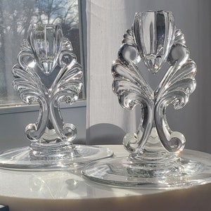 Può includere: Due portacandele in vetro trasparente con disegni decorativi a forma di fiore. I portacandele hanno una base circolare larga e una parte superiore alta e decorativa.