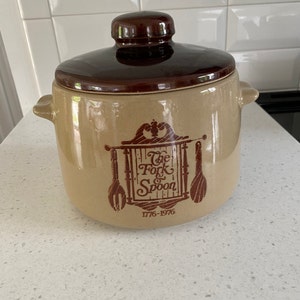 Może przedstawiać: Brązowo-beżowy ceramiczny garnek z pokrywką. Garnek ma brązową pokrywkę i uchwyty po bokach. Z przodu garnka widnieją słowa "The Fork & Spoon" oraz liczby "1776-1976" w kolorze brązowym.