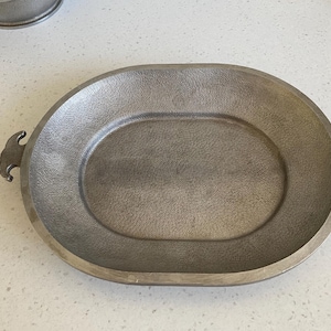 May include: Oval-shaped, pewter-colored serving platter with textured surface and decorative handles. The platter is made of metal and is designed for serving food. The handles are curved and add a decorative element to the piece.
