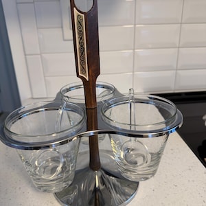 May include: A vintage condiment server with three clear glass cups held in a chrome-plated metal frame. The central handle is dark wood with a decorative inlay. The server sits on a round base.