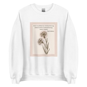 Könnte beinhalten: Weißer Pullover mit einem Zitat von Ray Bradbury: "Und plötzlich war alles, absolut alles, da." Das Zitat ist auf einem hellrosa Hintergrund mit schwarzem Rahmen und einer Zeichnung von zwei Blumen gedruckt.